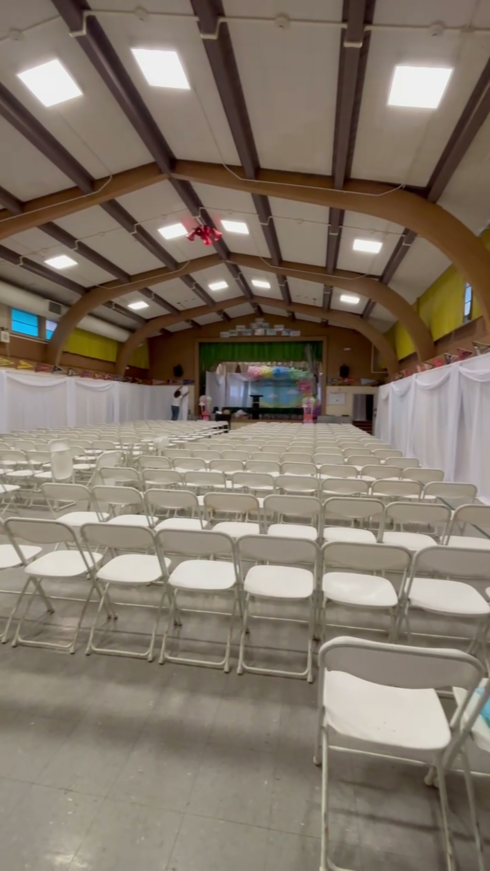 School gymnasium transformed into a graduation ceremony venue with white wall draping on both walls, rows of white folding chairs, and a colorful stage backdrop in Sacramento